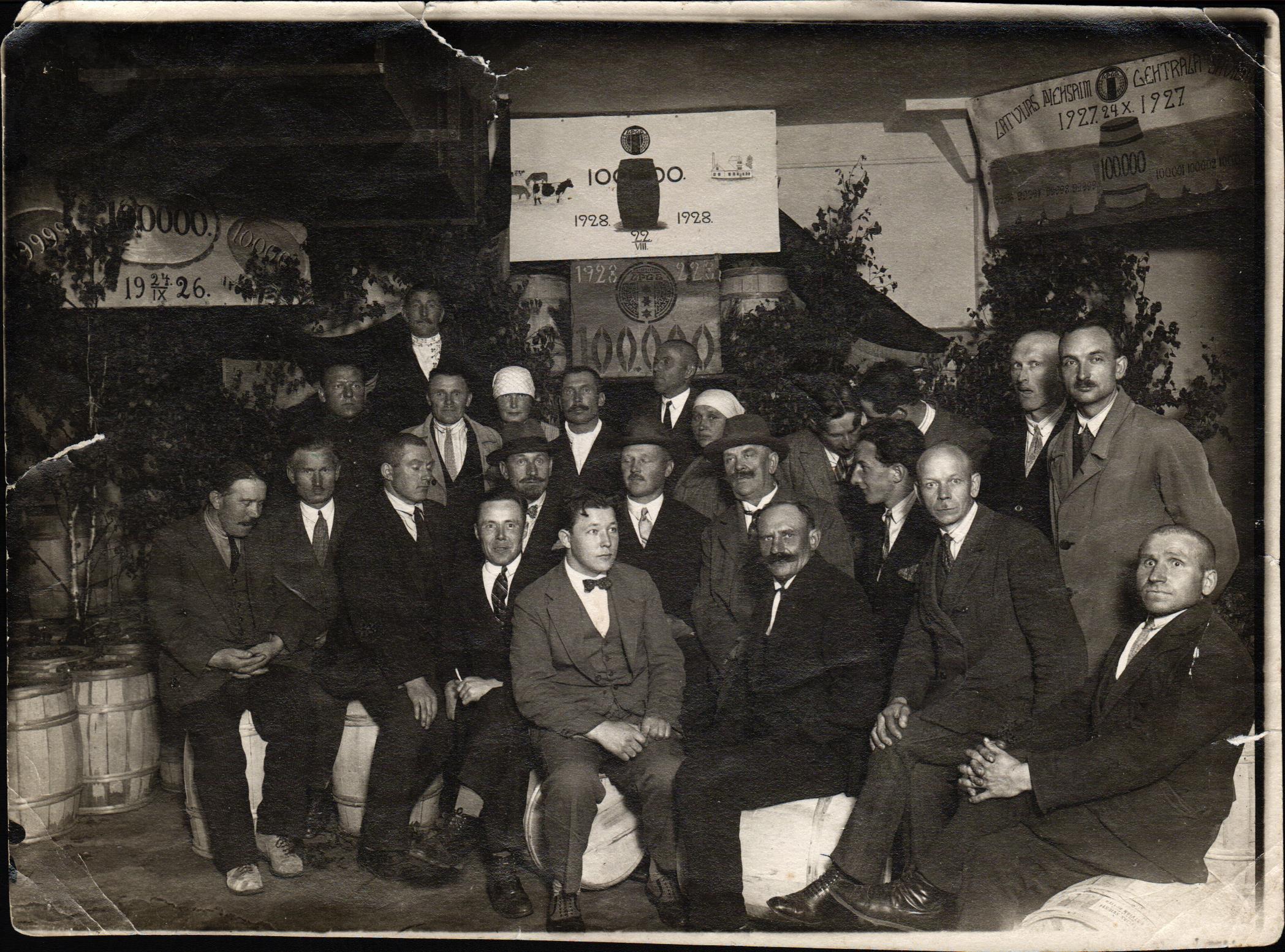 Latvijas Piensaimnieku Centrālās savienības darbinieki kādā svinīgā pasākumā ar sviesta muciņām fonā. No kreisās: 2. rindā 4. (ar hūti galvā) Teodors Kalniņš. A. Majevska foto. 20. gadsimta 30. gadi.